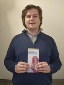 Ein Mann mit mittellangem braunem Haar, der einen blauen Pullover mit Reißverschluss über einem gestreiften Hemd trägt, steht in einem Innenraum vor einer beigen Wand. In der Hand hält er ein Flugblatt mit dem Foto eines lächelnden Mädchens und dem deutschen Text "Mädchen brauchen Ihre Unterstützung".