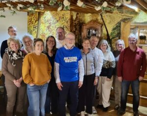 Eine Gruppe von zwölf Erwachsenen, Männern und Frauen, steht in einem Innenraum dicht beieinander und lächelt in die Kamera. Ein Mann im Vordergrund trägt ein blaues Hemd mit der Aufschrift "AKTIONS GRUPPE". Im Hintergrund sieht man rustikale Holzbalken, eine Wandmalerei aus Laub und eine warme, gemütliche Beleuchtung.